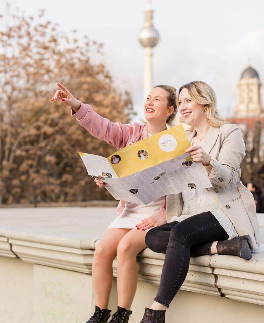 Hochzeitsfotograf Berlin Zwei Frauen sitzen auf einer Mauer vor dem Fernsehturm Berlin, lachen in die Ferne und zeigen auf etwas, dabei halten sie eine Karte fest